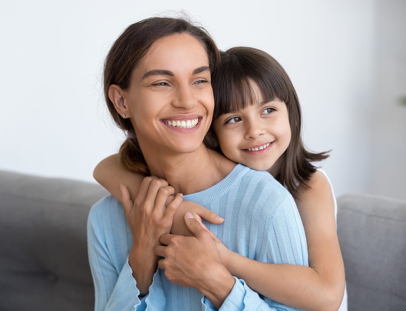 A smiling young girl hugs her mother from behind as they both sit together on a couch, sharing a joyful moment - Oro Valley Dentist A smiling young girl hugs her mother from behind as they both sit together on a couch, sharing a joyful moment - Oro Valley Dentist